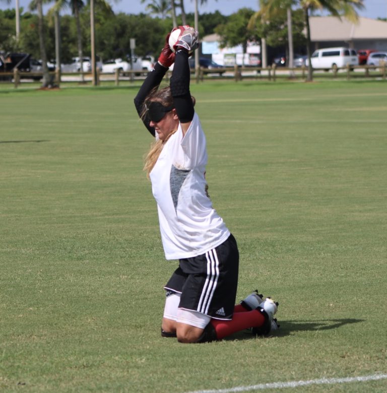 A blindfolded player rests on her knees while lifting the ball over her head with both hands.