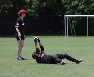 Jamie Sibson with the Austin Blackhawks laying on her back holding the ball up in the air making an out with spotter Molly Fleming looking on.