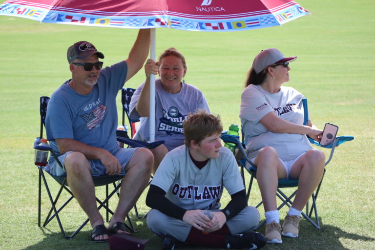 A BCS Outlaw's player rests in the shade of an umbrella held up by a few fans.