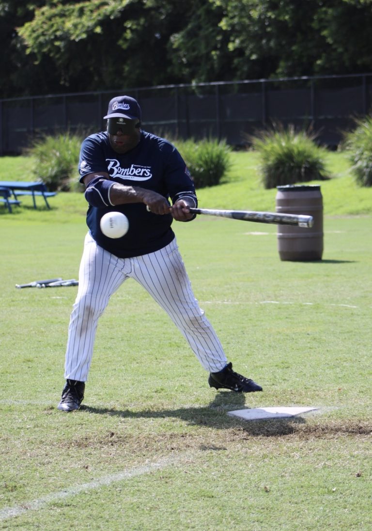 A Long Island Bombers player swings the bat as a ball flies towards him.